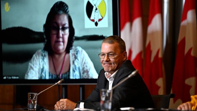 A man speaks at a press conference with Canadian flags and a television behind him.