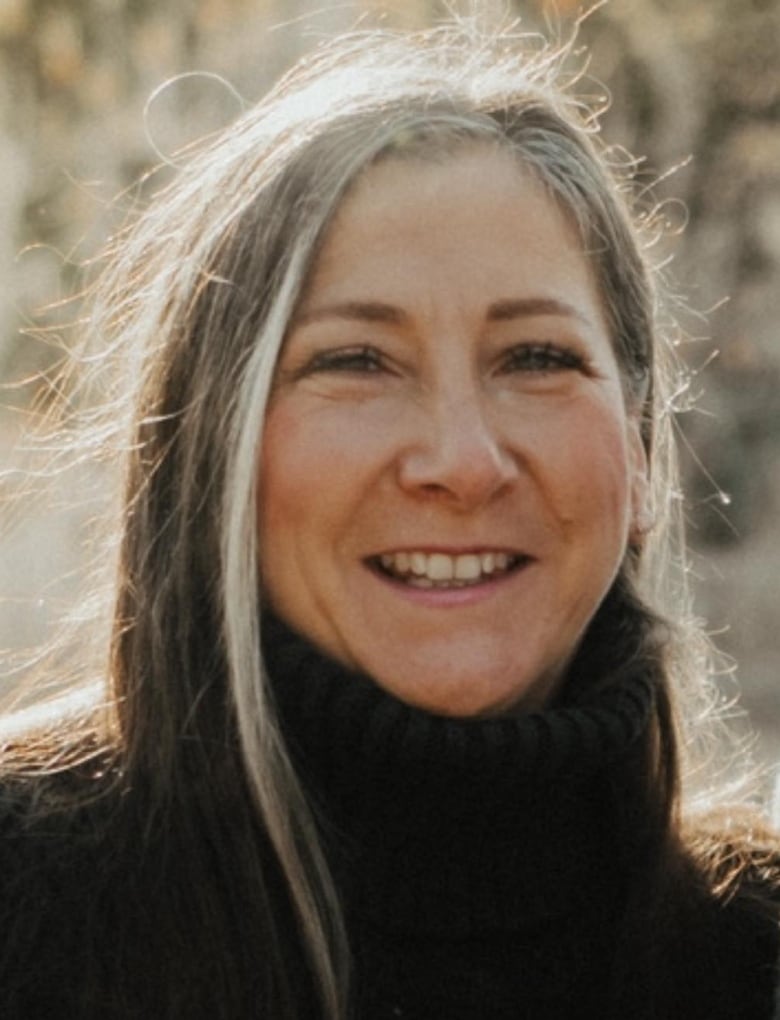 A woman outdoors, smiling at the camera. She's wearing a black turtleneck and has dark hair with a grey streak in the front.