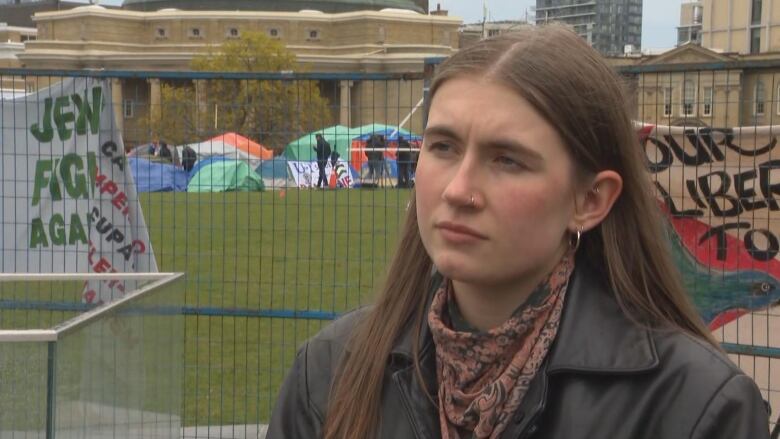 A woman looks off camera. A fence, tents and signage are seen in the background.