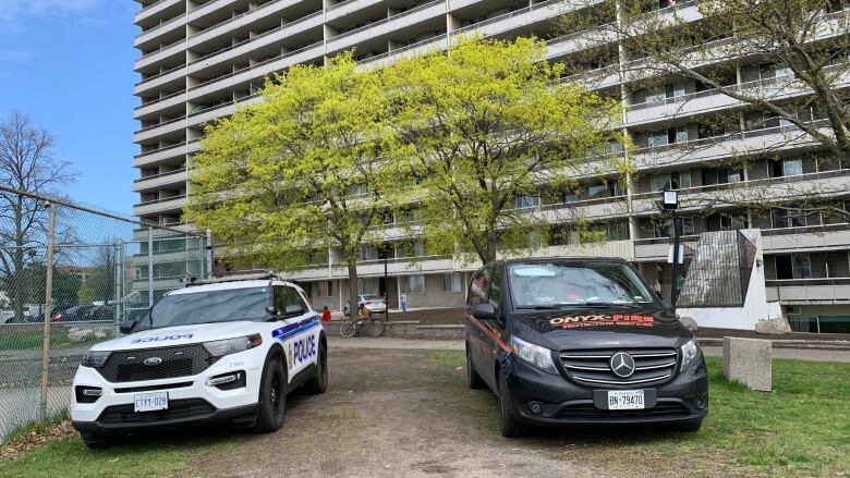 Two vehicles, one of which is a police cruiser, is parked in front of a highrise. 