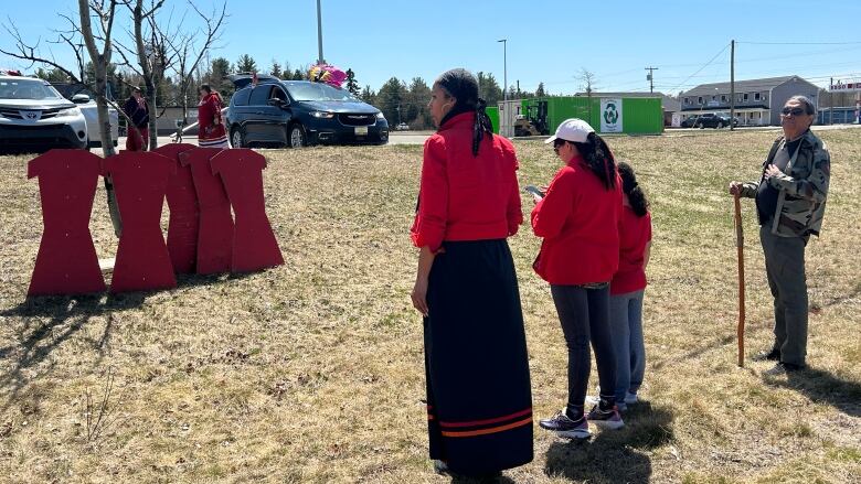 People stand and look at four cutout red wooden dresses standing against a tree.