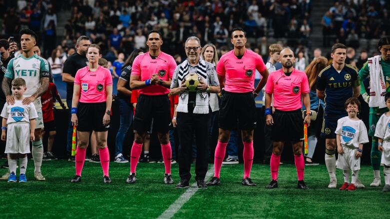 A man holds up a yellowed soccer ball as he is flanked by referees, players and mascots.