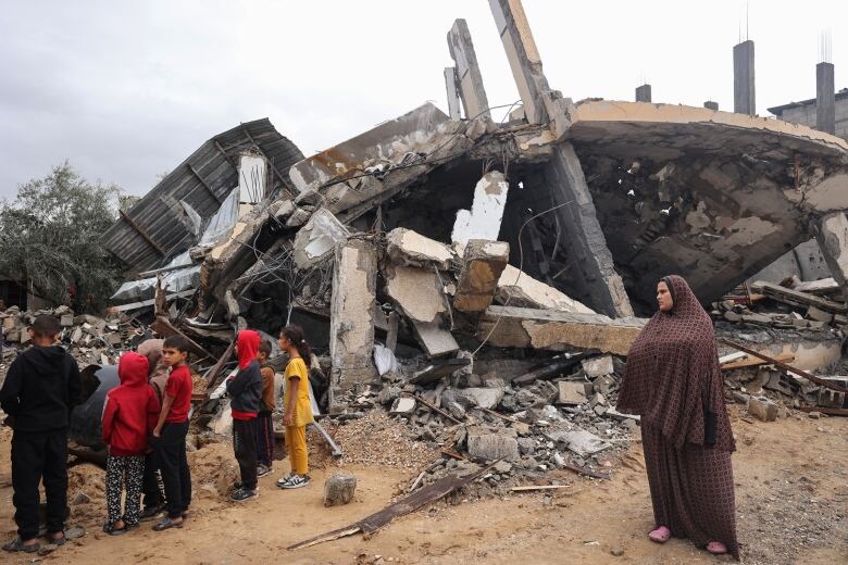 A woman wearing a head covering and several children are shown near a large damaged concrete structure, with stone debris scattered on the ground below.