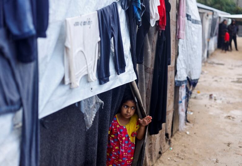 A small child peers out of a tent covered with hanging clothers in a makeshift camp.