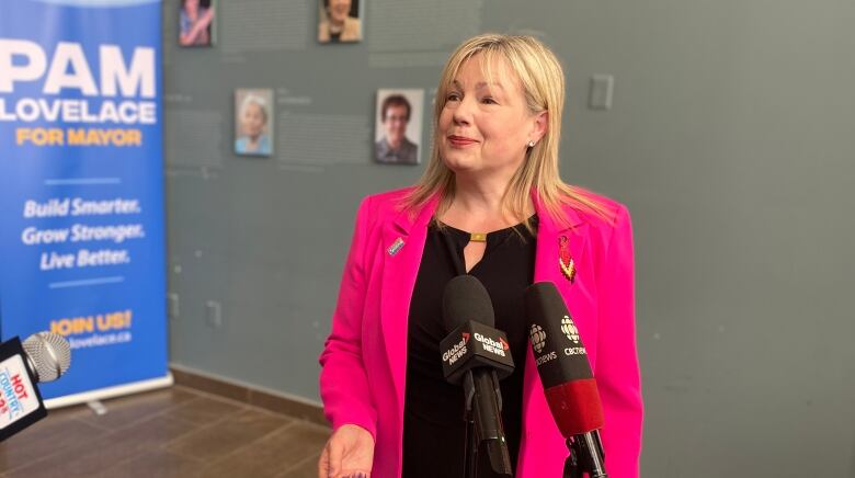 A woman in a black dress and a bright pink blazer stands in front of two microphones in a grey room.