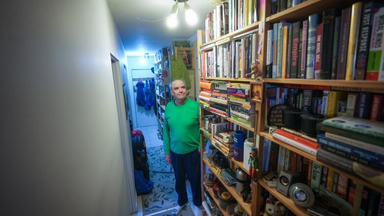 A man stands next to a towering shelf filled with books covering his entire hallway.