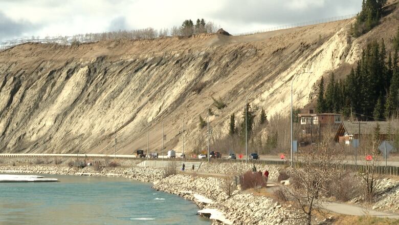 Vehicles are seen from afar on a road running along the base of a steep slope.