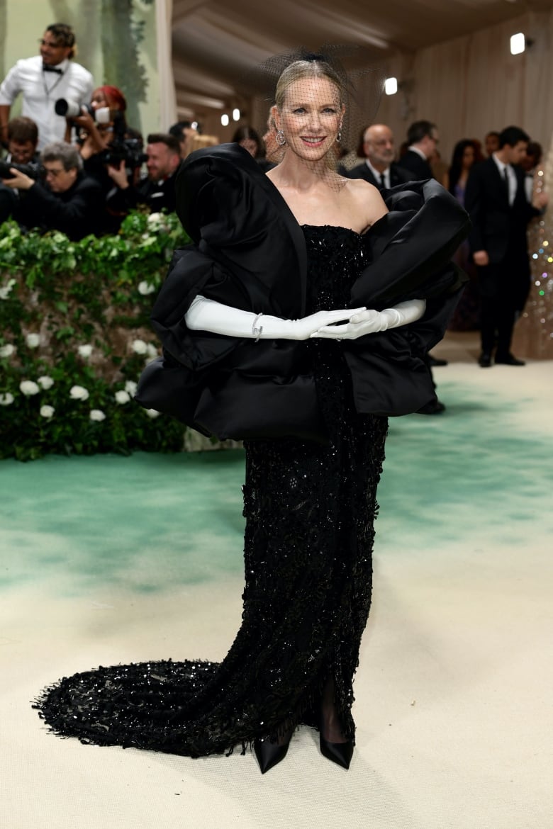 Naomi Watts on the Met Gala red carpet wearing a beaded black gown, puffy cape and white gloves.