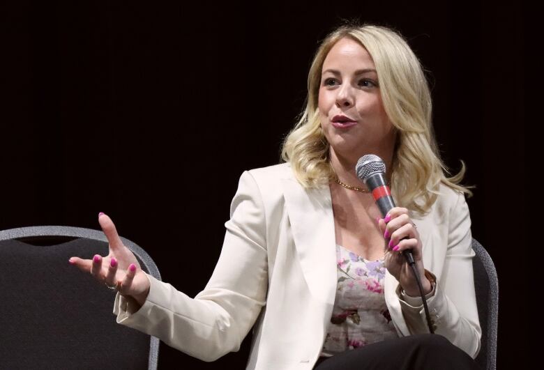 A woman with a cream suit jacket, print blouse, blonde hair and bright pink nails holds a microphone while speaking and gesturing with her hand.