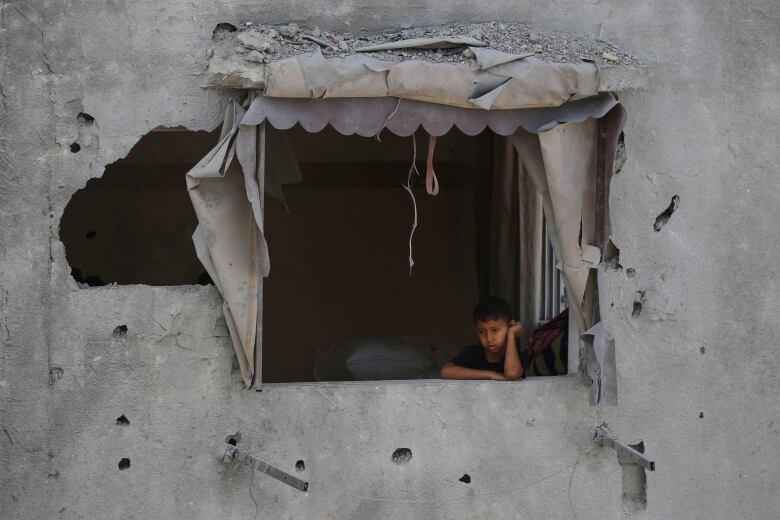 A small boy rests his head against his arm while sitting at a ledge without a window of a damaged concrete structure.