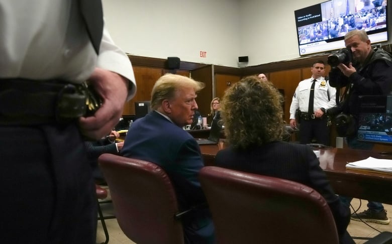 A defendant in a blue suit leans over to speak to his lawyer from a table in a courtroom.