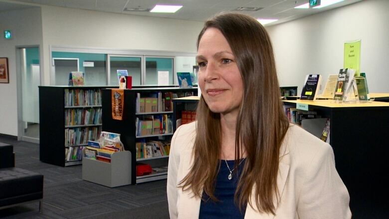 A woman with long brown hair stands in front of several book shelves. She wears a white blazer. 