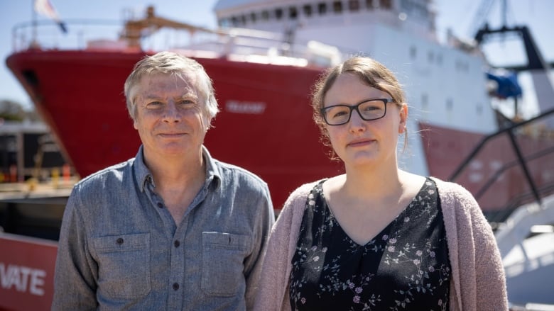 A man and a woman stand in front of a DFO boat. The man is wearing a blue chambray shirt, while the woman wears a purple cardigan, and a blouse with flowers on it. 