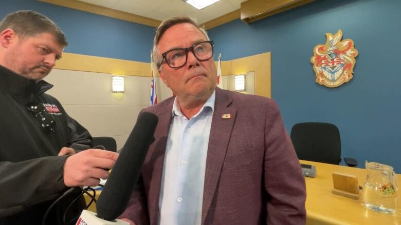 A white man wearing a purple suit jacket answers a question in a city hall.