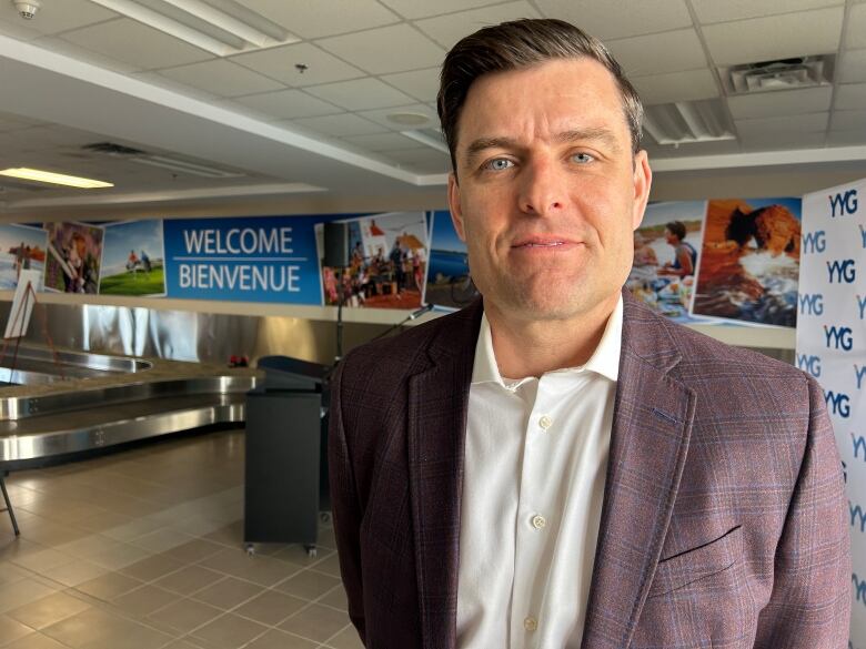 A man with short hair and a red checked blazer stands in the baggage and arrivals area of an airport terminal.