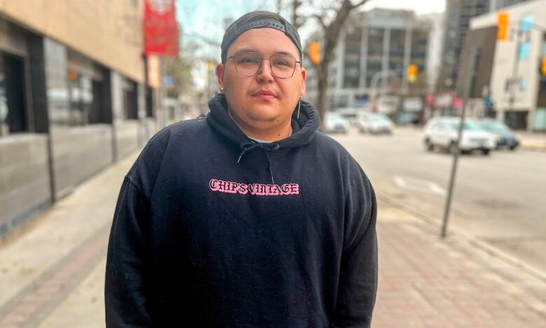 A person wears a black hooded sweatshirt, backwards ball cap and glasses. They stand on the sidewalk of a downtown street. 