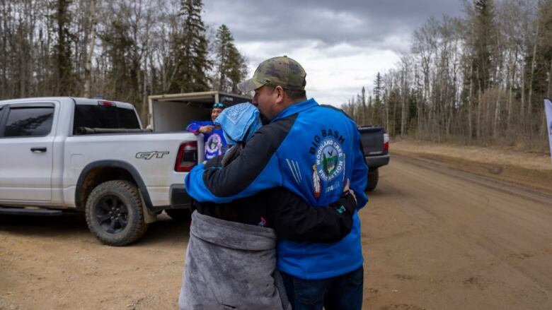 Two people hug on the side of a dirt road. A truck is in the background.