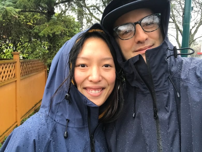 A woman and a man, each wearing dark blue rain ponchos, stand in the rain.