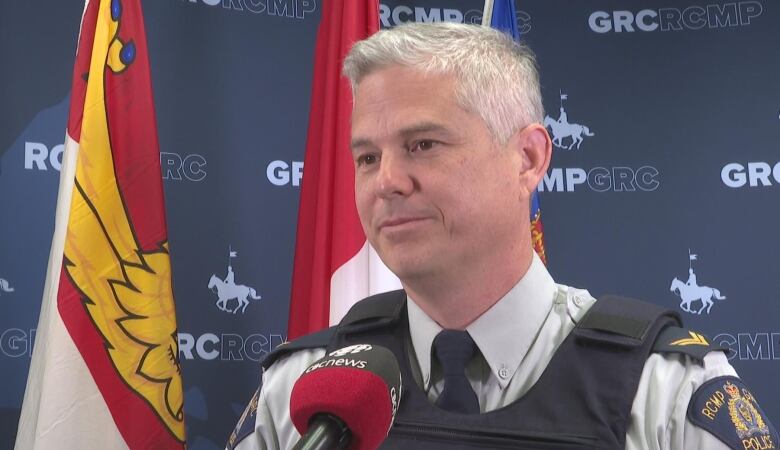 A police officer with grey hair, wearing a bulletproof vest in front of a Mountie-themed official backdrop. 