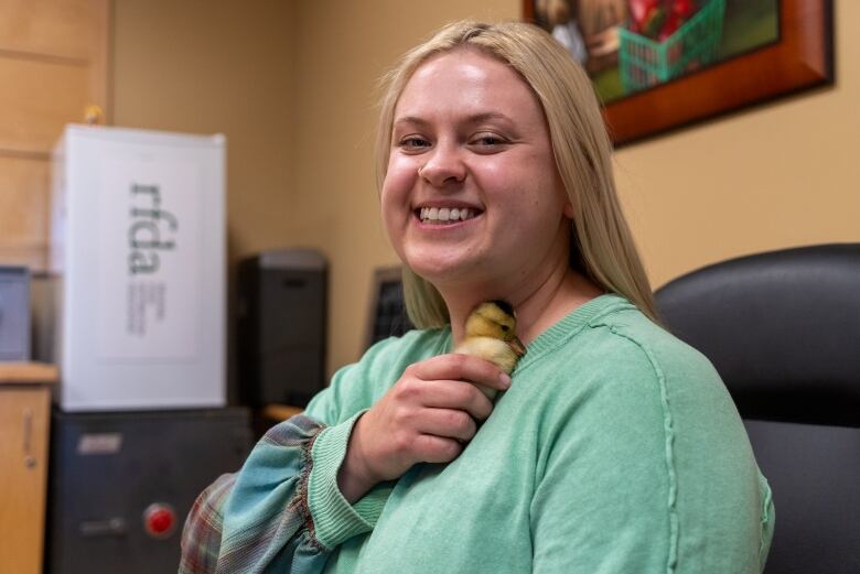 A person with long blonde hair and a green sweater holds a baby duck in their hand. The duck is nuzzled into their neck.