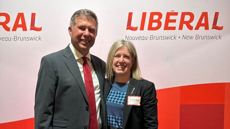 A smiling man dressed in a suit and tie, standing with his arm around a smiling woman, wearing a dress and blazer, with a red and white New Brunswick Liberal Party sign behind them.