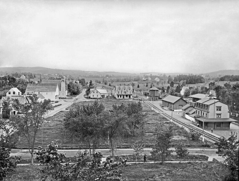 A black and white aerial shot over a town square with a railway station on the left and green space in the centre. Some wooden homes and larger buildings around the sides.