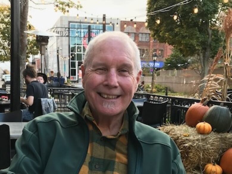 A man with gray hair and wearing a green jacket and green and yellow plaid shirt smiles at the camera. In the background is a streetscape with decorative white lights on a restaurant patio and fall decorations including hay bales and gourds.