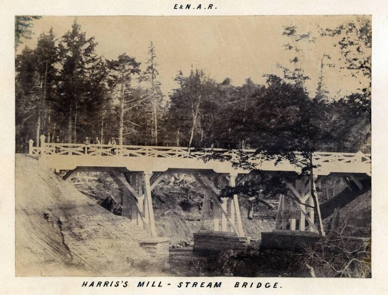 A white wooden beam bridge on top of stone piers across a narrow stream. Side view. A few people leaning over the railing towards the camera. Shot from roughly 100 metres away. Words in margin: Harris's Mill Stream Bridge.