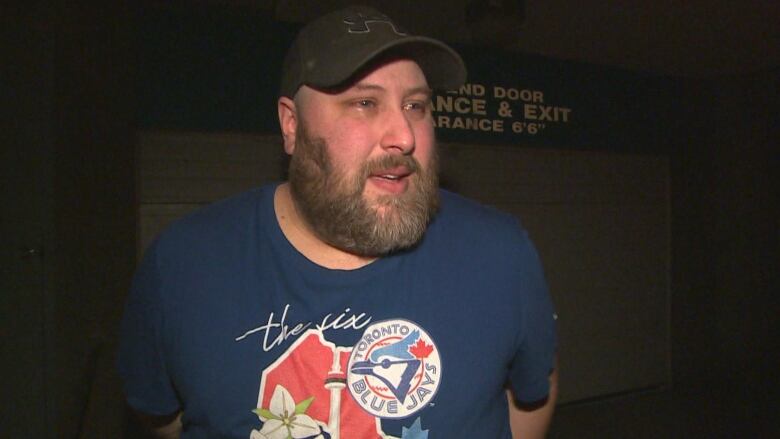 A man in a baseball cap, blue T-shirt and beard has teary eyes and looks to the right of the image.