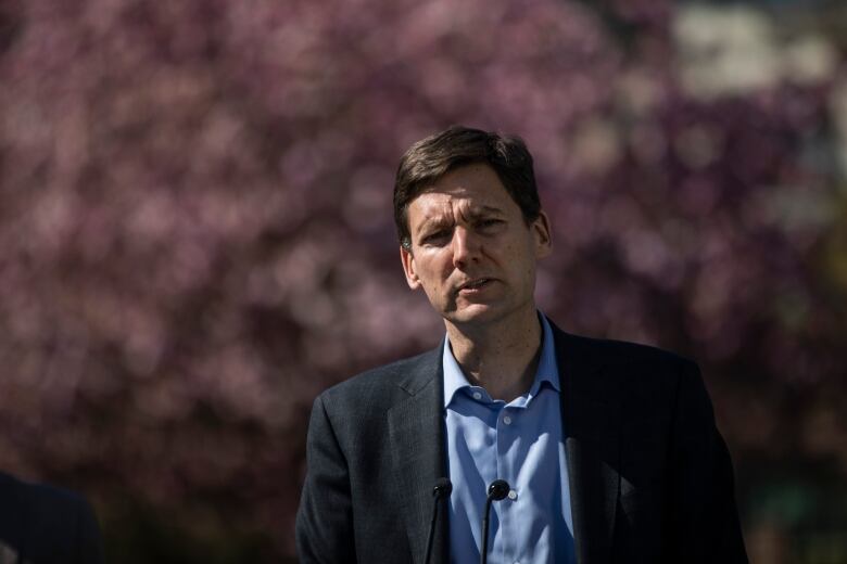 A tall white man speaks in front of a cherry tree in full bloom.