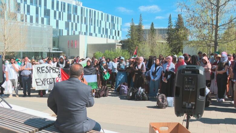 a  group of people stand in a crowd. some hold palestinian flags. a man speaks into a microphone with a speaker facing the crowd.