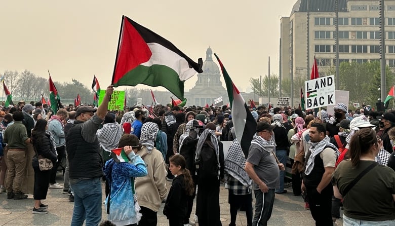 A crowd of people gather on the Alberta Legislature grounds. The sky is clouded with wildfire smoke. Some Palestinian flags are being waved. The legislature stands hazy in the background.