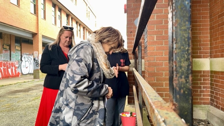 One woman, and two others in the background, stand near a metal gate that encloses a window well of a red brick building. She gazes down into the well.