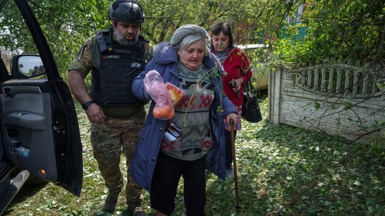 An elderly woman in mauve coat carrying a bag of oranges and using a cane walks with a woman in red and a medic wearing a helmet, camouflage and body armour.