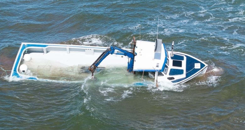 A drone view of a half-submerged white and blue fishing boat near a sandbar, being hit by waves. 