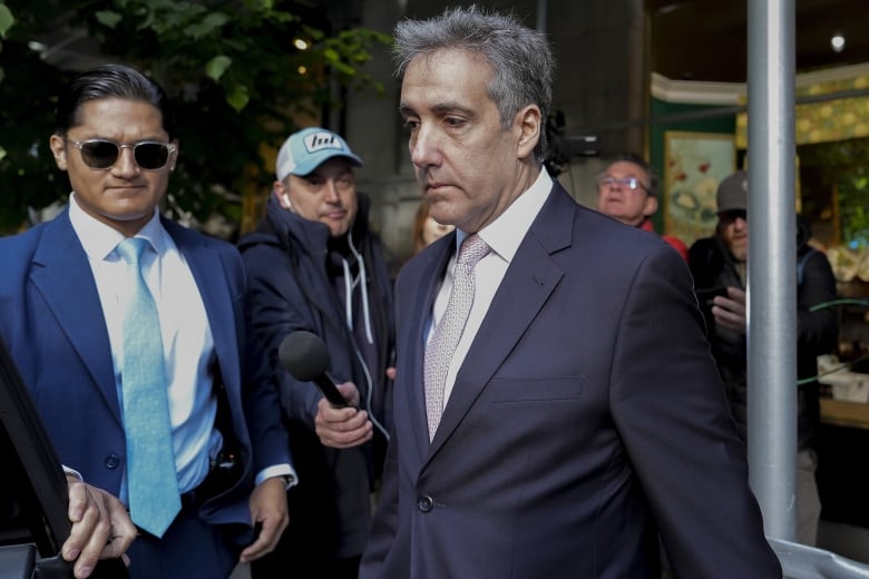 A grey-haired man in a navy suit walks outside surrounded by other people.