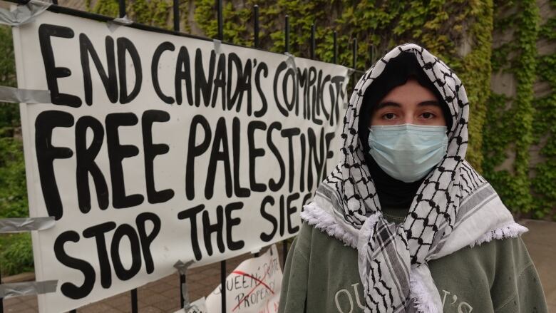 A woman wearing a keffiyeh scarf and a blue surgical mask stands in front of a large poster that states 