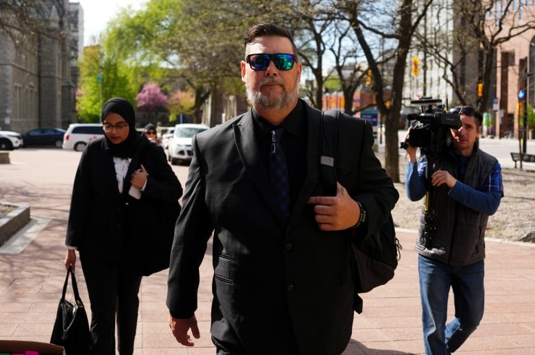 A man walks outside in a suit and sunglasses.