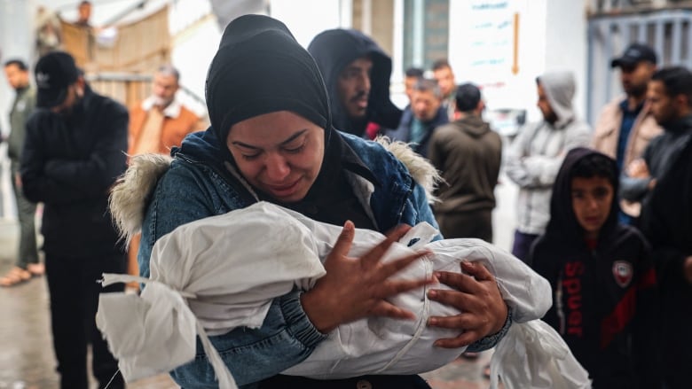 A woman with a black scarf over her head and wearing a denim jacket holds a child, shrouded in a small with body bag, in her arms.