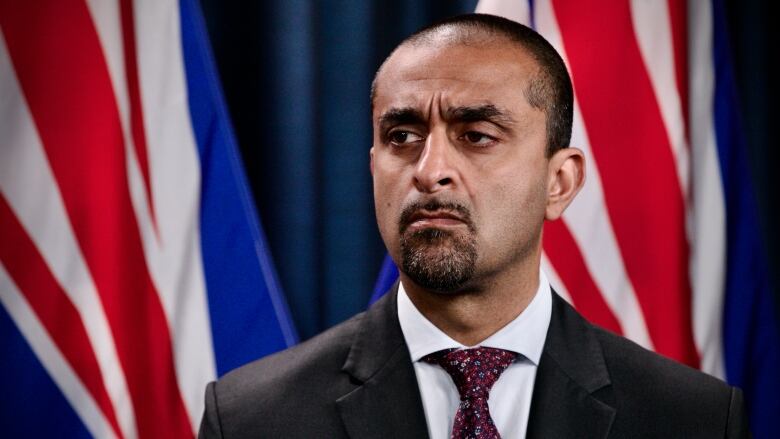 A South Asian man looks to the side, while in front of B.C. flags.