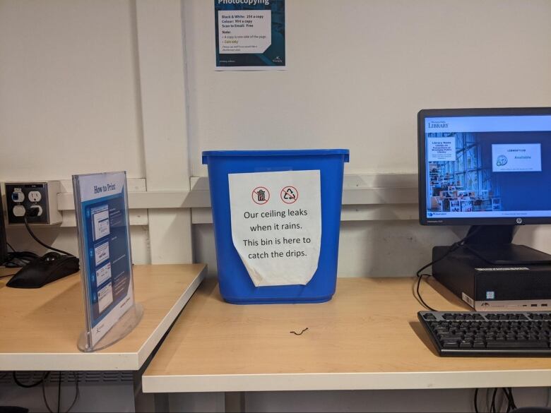 A blue recycling bin is shown on a desk next to a desktop computer. A sign taped onto the bin reads, 'Our ceiling leaks when it rains. This bin is here to catch the drips.'