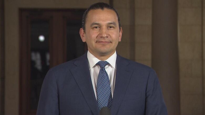 A man wearing a navy blue suit and tie is pictured from the waist up.