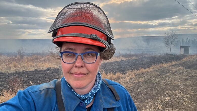 Woman in orange hard hat and blue overalls stands in a scorched out field on a wildfire crew. 