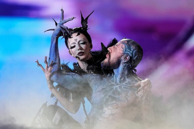 Two people in elaborate costumes and makeup perform on a smoke filled stage. 