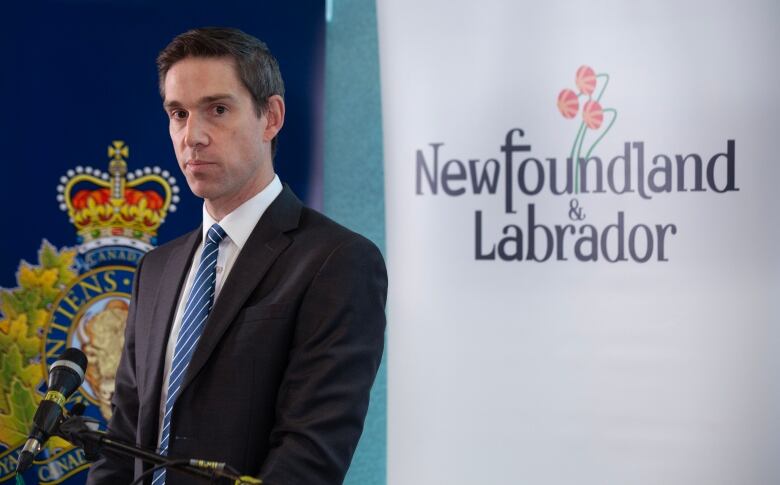 A man wearing a suit stands silently at a lectern, in front of posters showing the Royal Newfoundland Constabulary shield and the words 