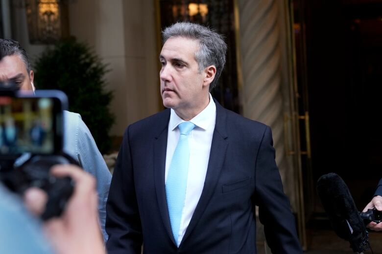 A man in a black suit and blue tie leaves his apartment building. Camera crews surround him.