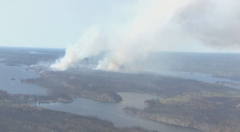 Columns of wildfire smoke rise above forest and lake in the distance of an aerial image.