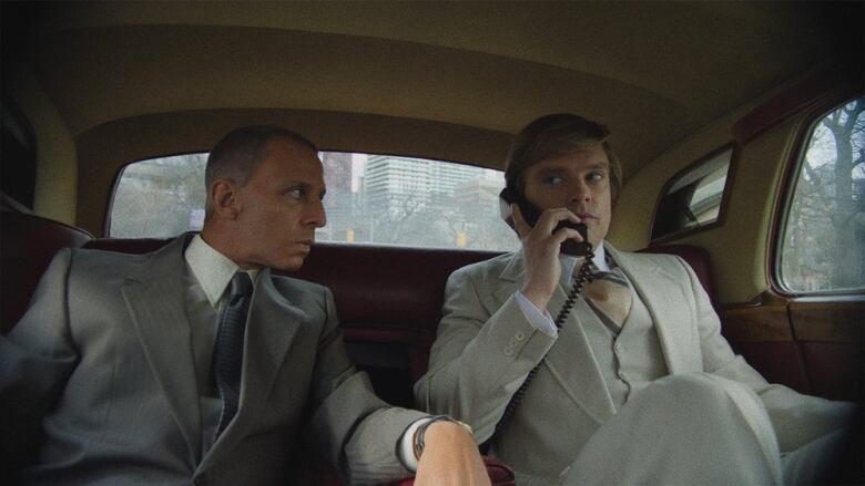 Two men in suits sit in the back seat of a car. The man on the right wears a cream coloured suit and speaks on a car phone. 