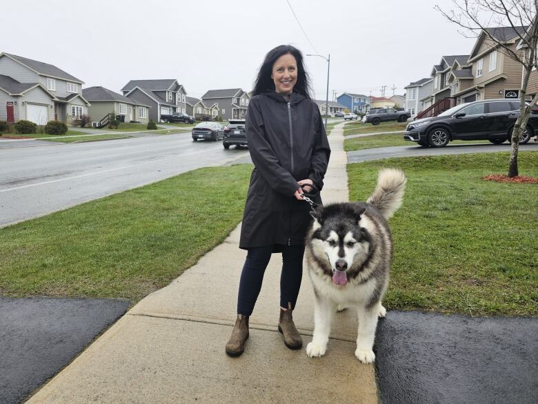 Woman in blue rain jacket standing on sidewalk holding leash to a large dog.