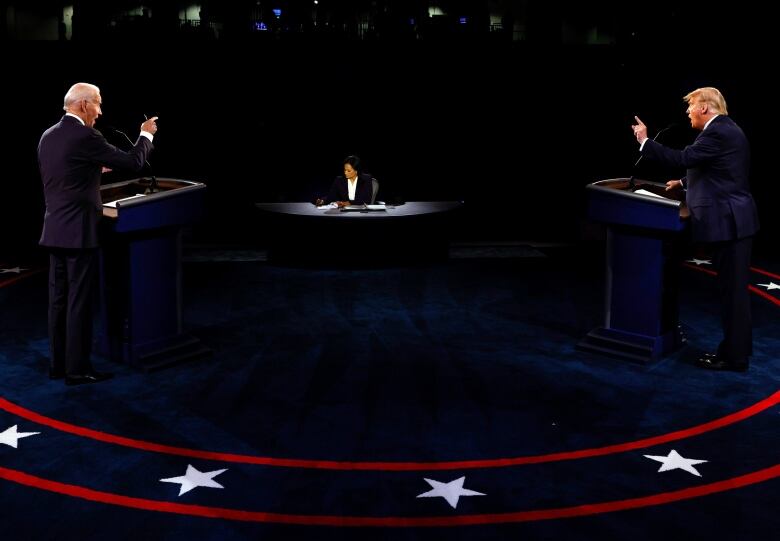 Two men, with their backs partially turned, stand behind podiums with a woman seated at a desk in the background in between the podiums. 